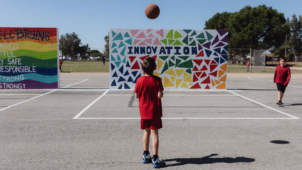 Boys play with a ball in a school playground. Behind them is a colorful mural. 