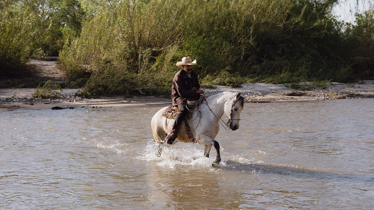 A man in a cowboy hat forges a river on horseback. 