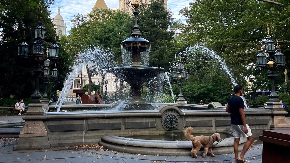 A person and his dog around the Jacob Wrey Mould Fountain in New York City's City Hall Park in Lower Manhattan with the spire of City Hall visible in the distance.