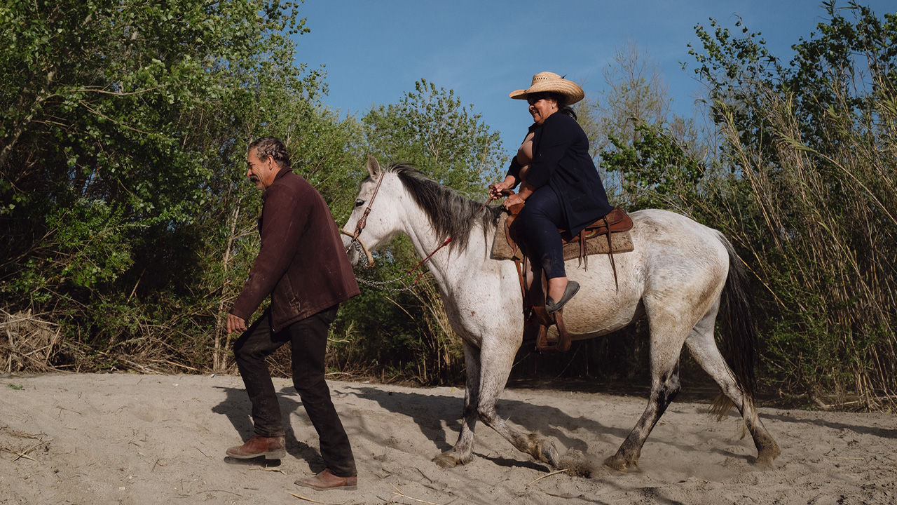 A man guides a woman riding on horseback along a sandy riverbank. 