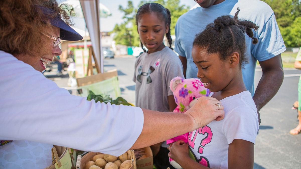 A woman reaches out to a young girl and her sister at a farmers market.