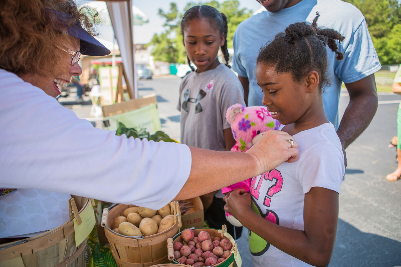 A woman reaches out to a young girl and her sister at a farmers market.