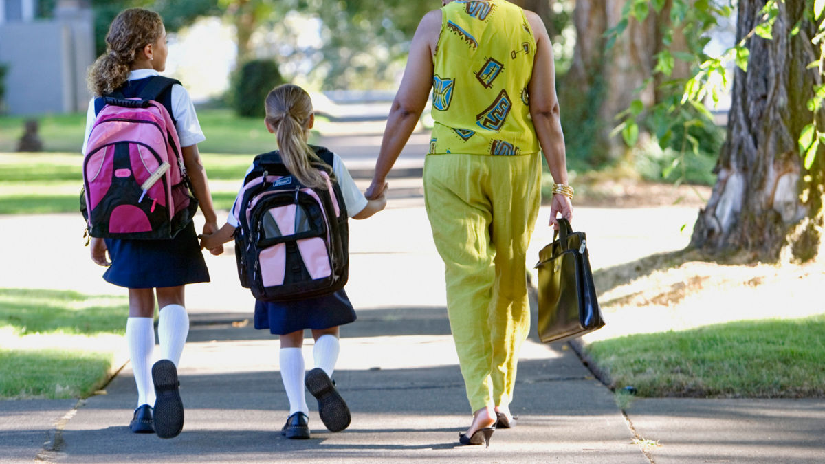 Rear view of mother with daughters walking down sidewalk. Daughters have backpacks, mother has briefcase.