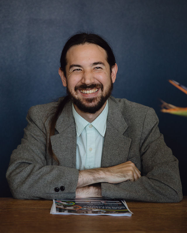 A community leader sits at a table against a blue wall, smiling.