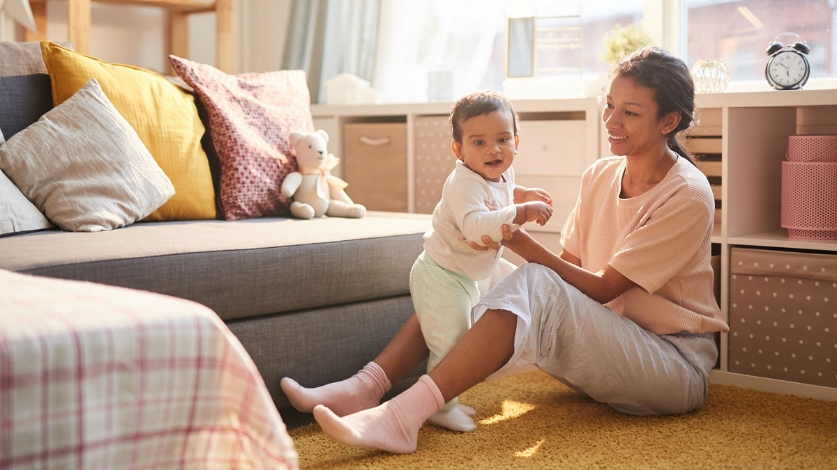 A mother sits on the floor with her baby, supporting him while he stands.