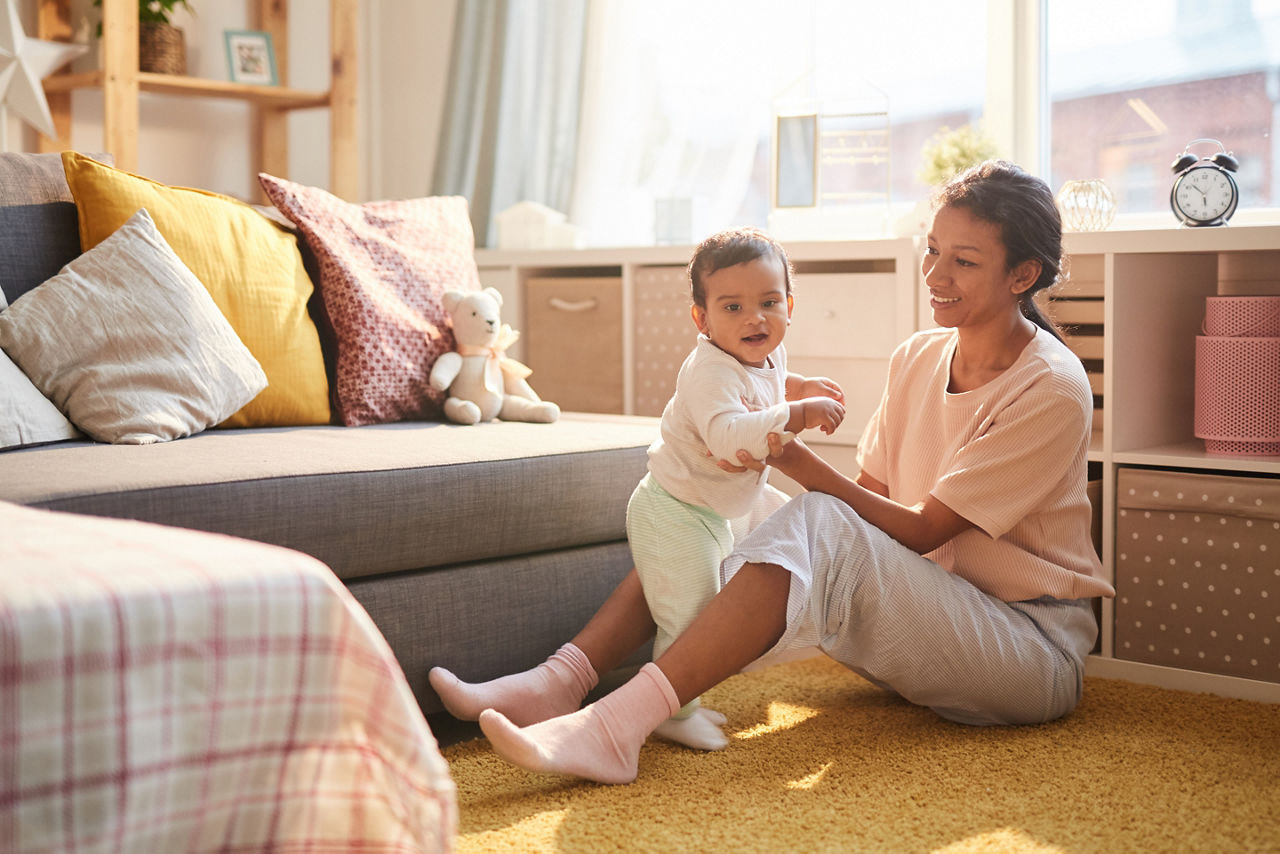 A mother sits on the floor with her baby, supporting him while he stands.