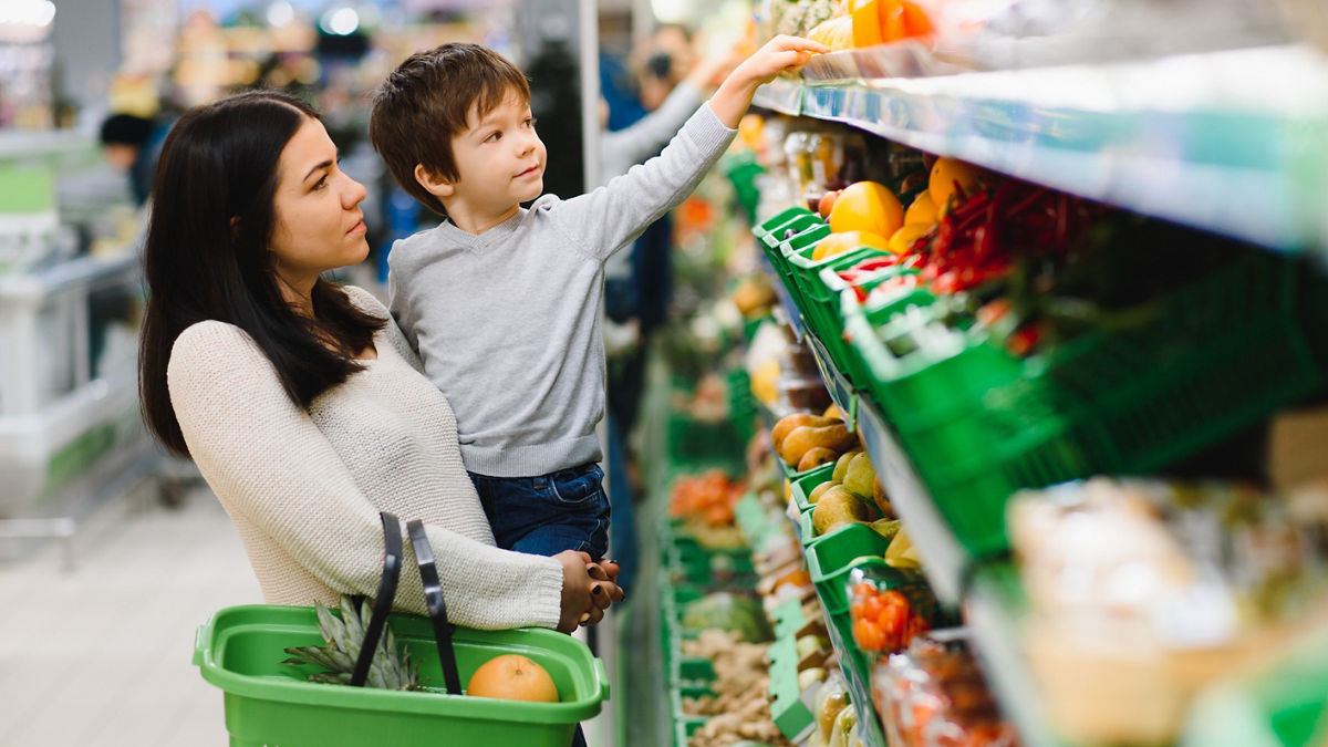 A mother holding her son up to a high shelf at the grocery store.