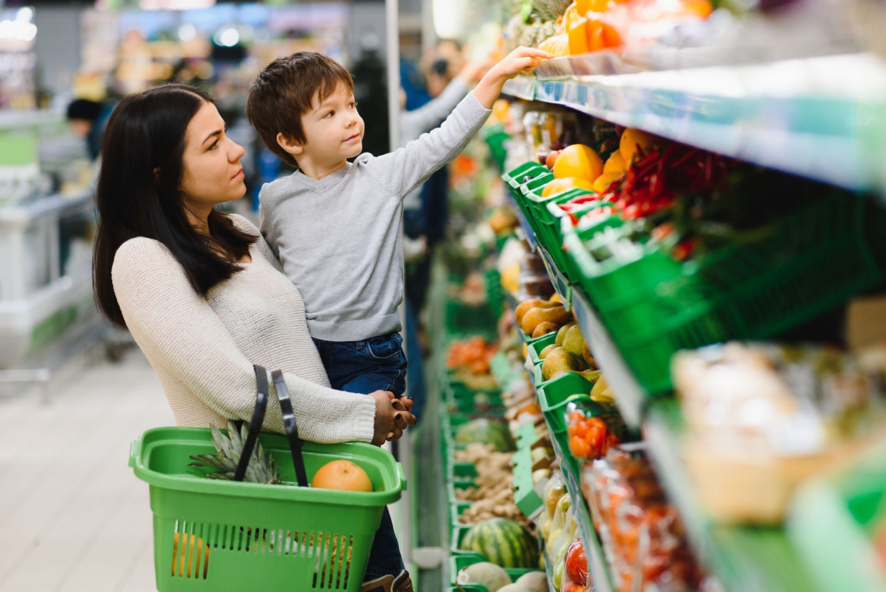 A mother holding her son up to a high shelf at the grocery store.