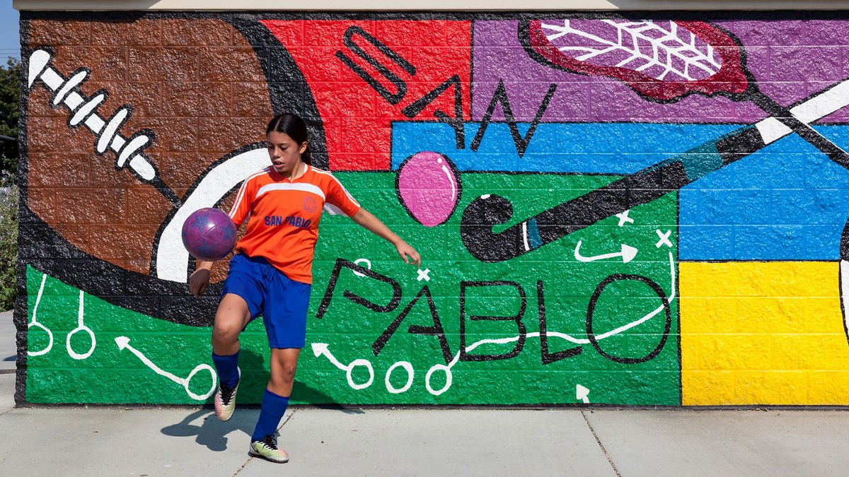 A youth soccer player juggles a ball in front of a mural highlighting sports equipment.