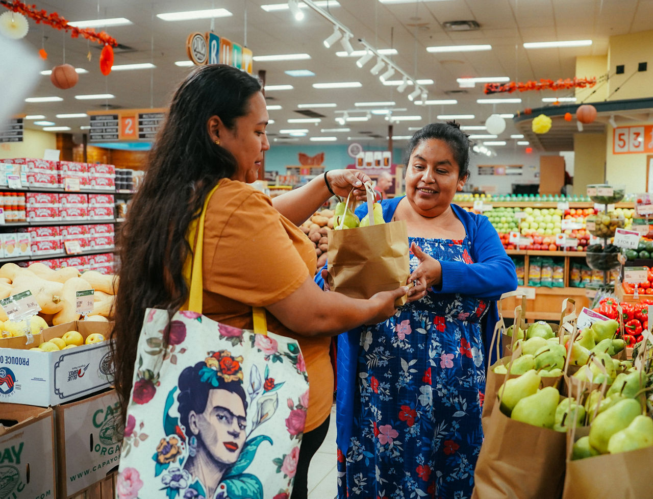 A woman hands a bag of fruit to another woman in an indoor market.