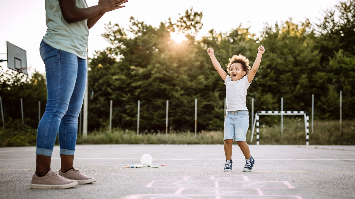 A young child playing hopscotch in a school yard with an adult.
