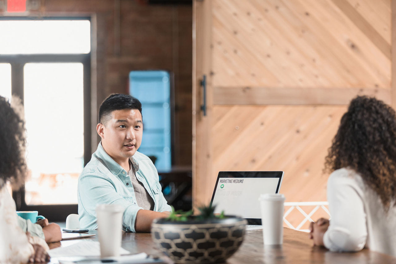 A male marketing executive discusses marketing strategy with a group of colleagues. He is using a laptop during the presentation.