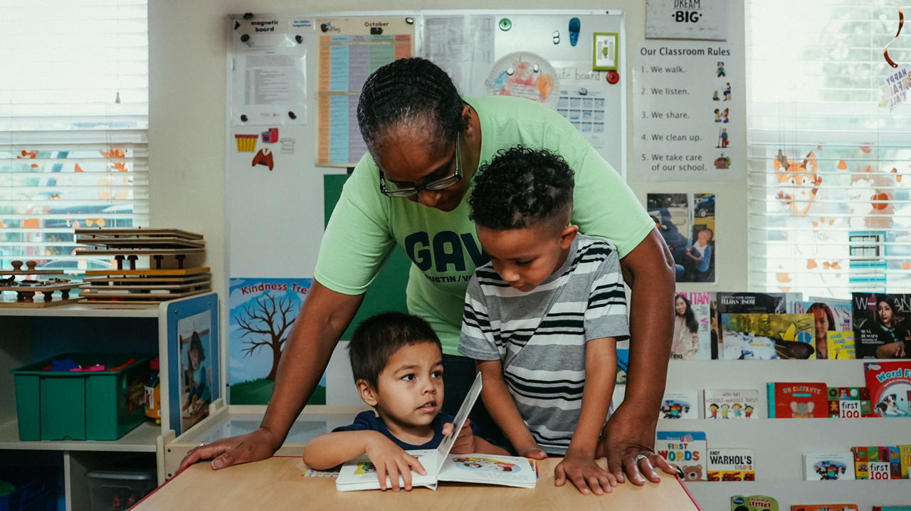 A person standing over two children reading a book.