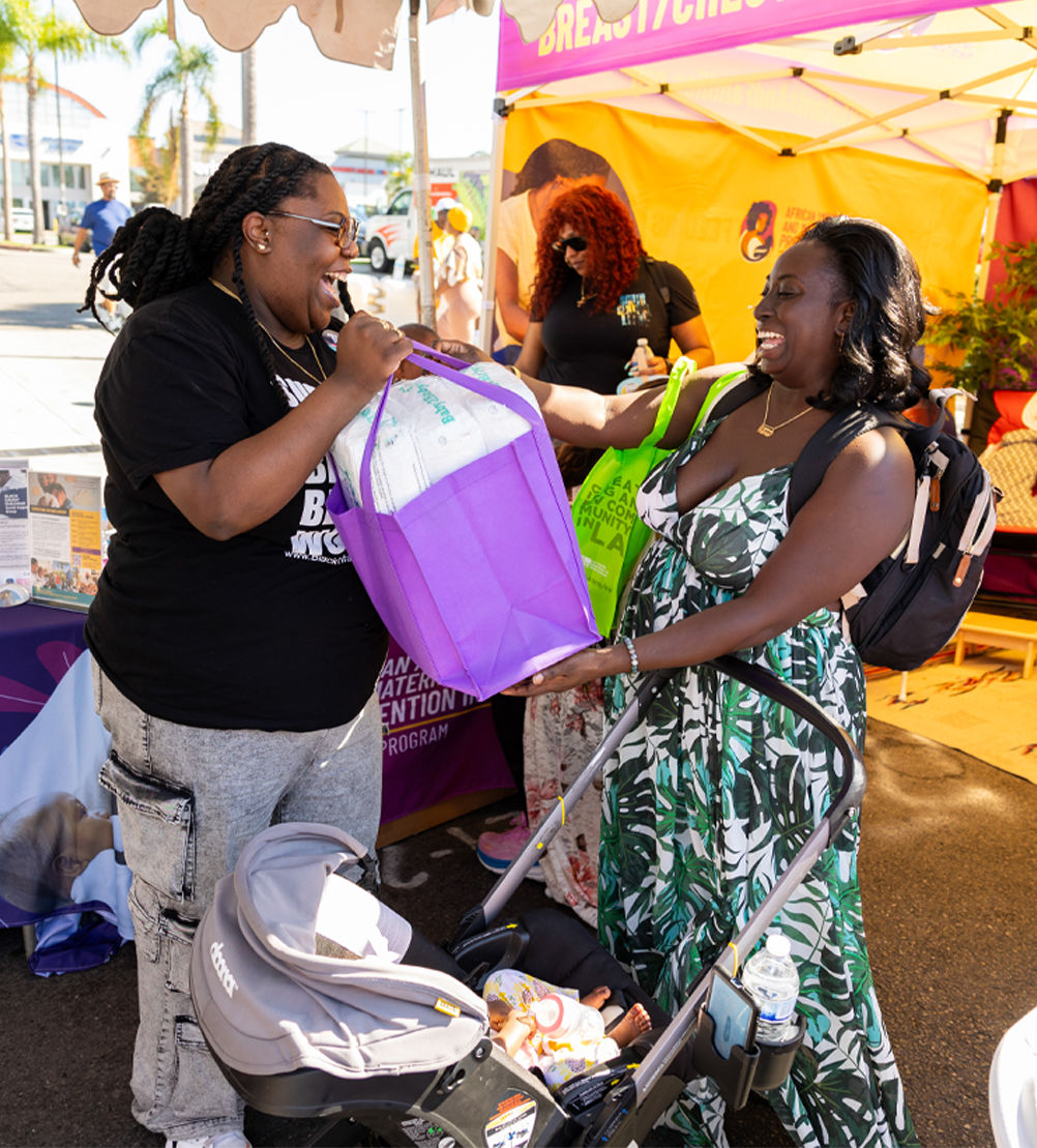 A person handing a purple shopping tote full of diapers to another person who is pushing a baby stroller.