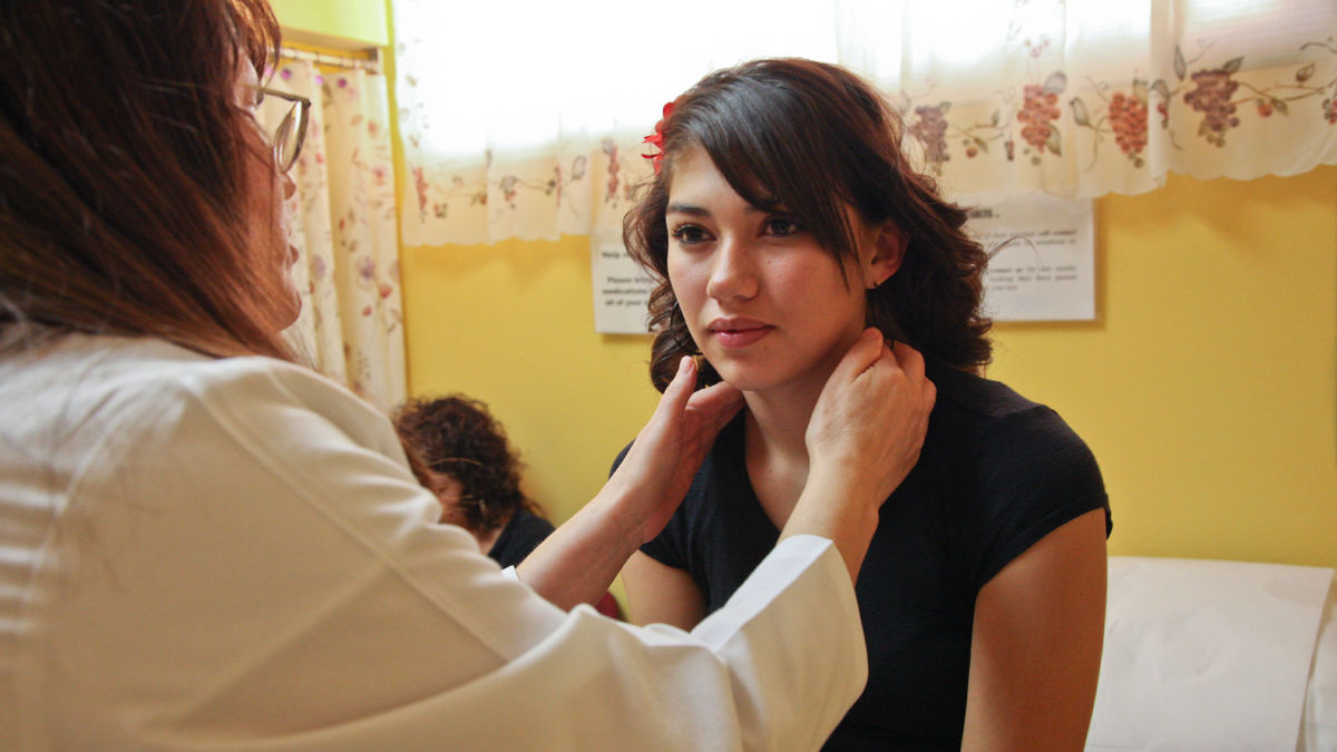 A doctor examines a teenage patient, her mother sits in the background, Hidalgo Medical Services-Bayard Community Health Center, Bayard, New Mexico. Project ECHO.