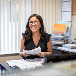 A happy, young woman sitting at a desk holding paperwork.