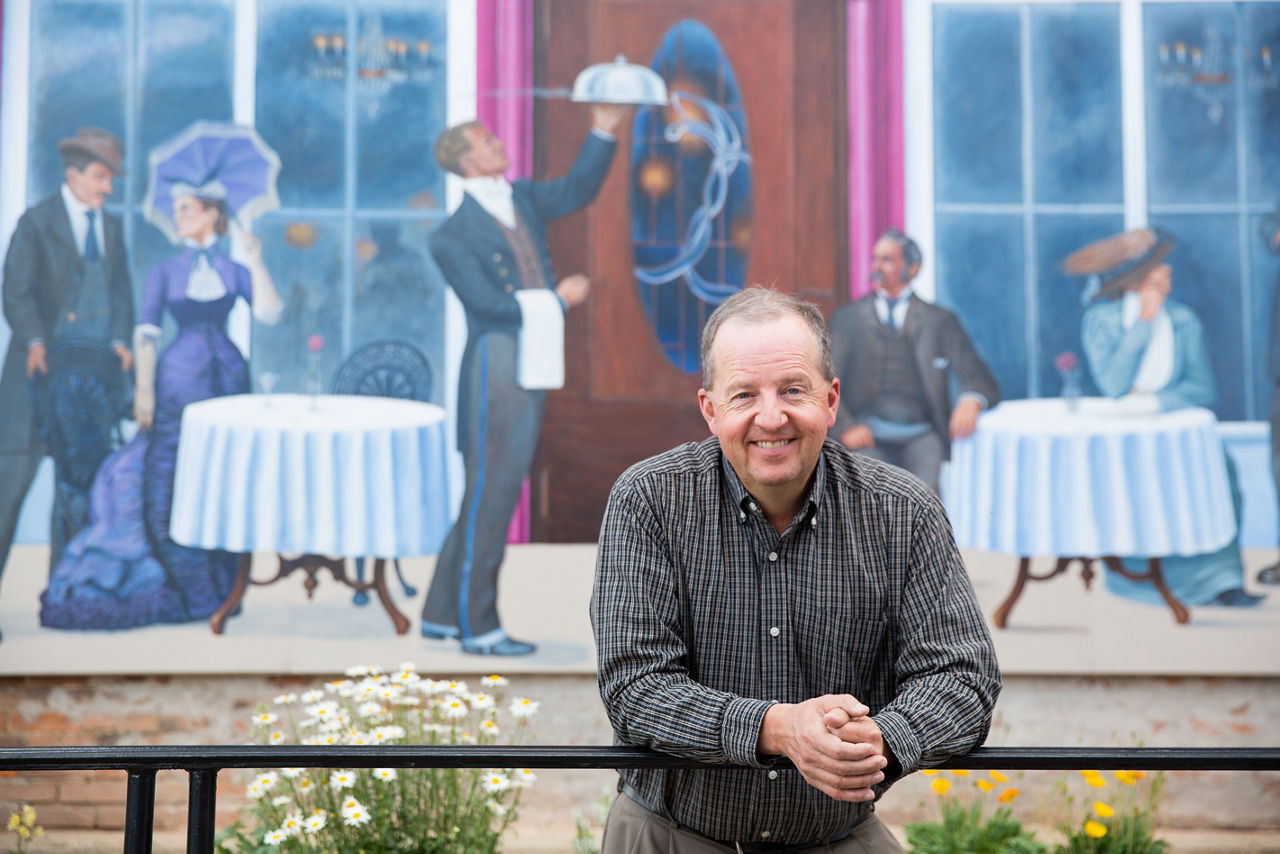 A man leaning on a railing, with a mural of people dining in the background.