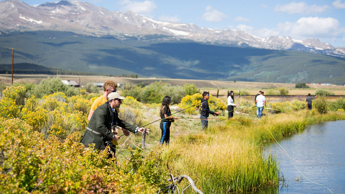 High school students fly fishing at a lake with a scenic, mountainous backdrop.