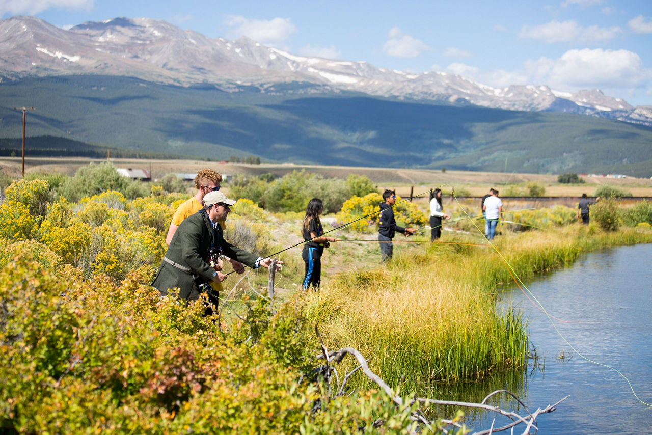 High school students fly fishing at a lake with a scenic, mountainous backdrop.