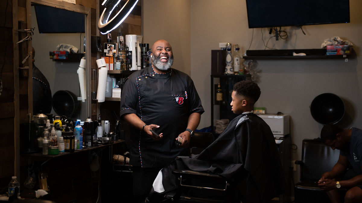 A barber smiles as he prepares to cut a young man’s hair.