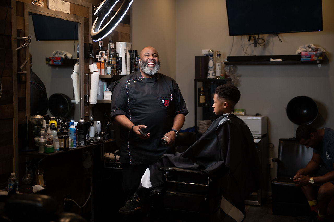 A barber smiles as he prepares to cut a young man’s hair.