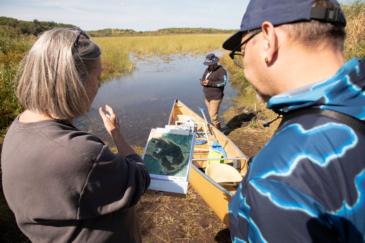 Two people are talking to each other while facing a lake. A third person is in the background looking at the water.