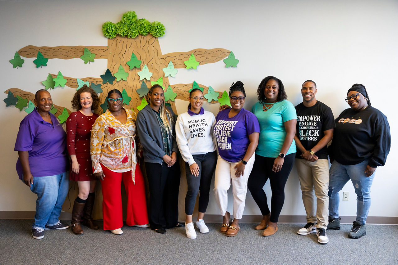 Ten people stand together in front of a large construction paper image of a tree.