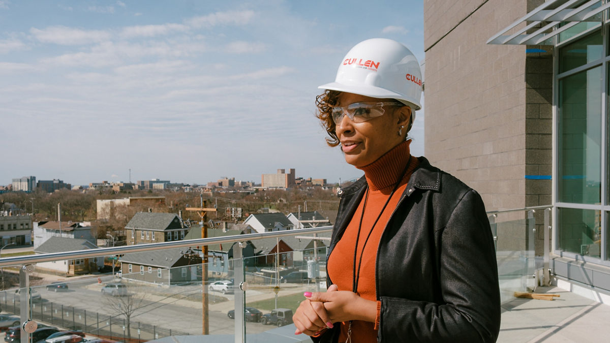 Woman wearing construction hard hat stands on a balcony looking over the surrounding neighborhood.