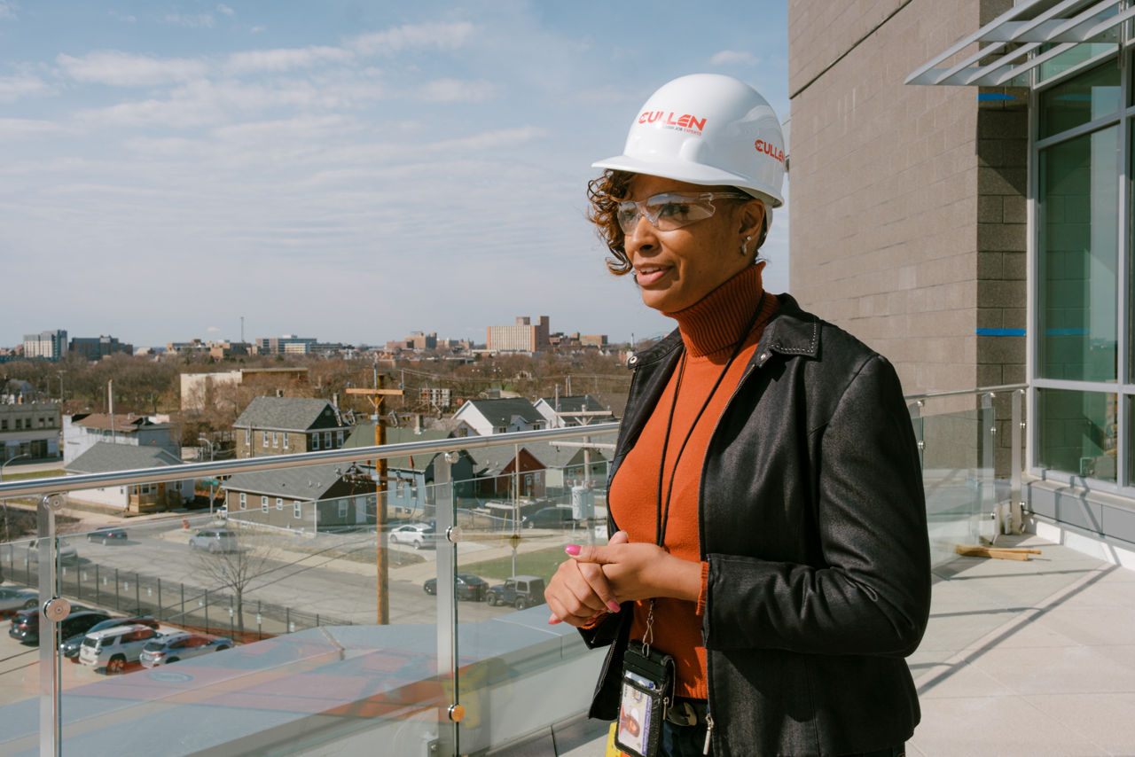Woman wearing construction hard hat stands on a balcony looking over the surrounding neighborhood.