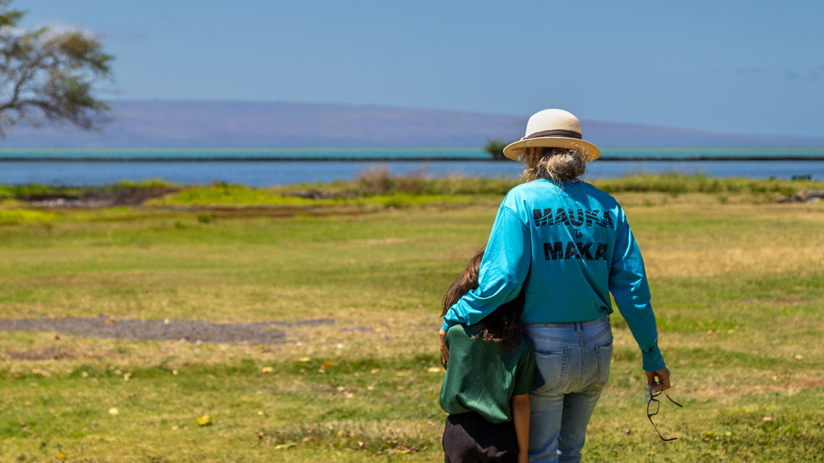 A woman in a straw hat stands near a body of water with her granddaughter.
