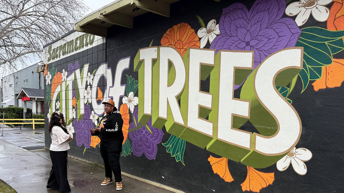 Two young people talk to each other on the sidewalk in front of a mural.