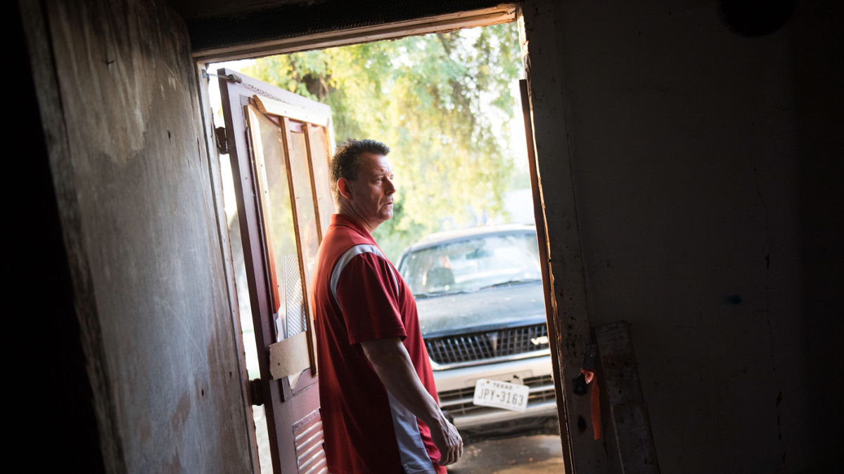A man stands in the doorway of a garage with a truck parked in the driveway.