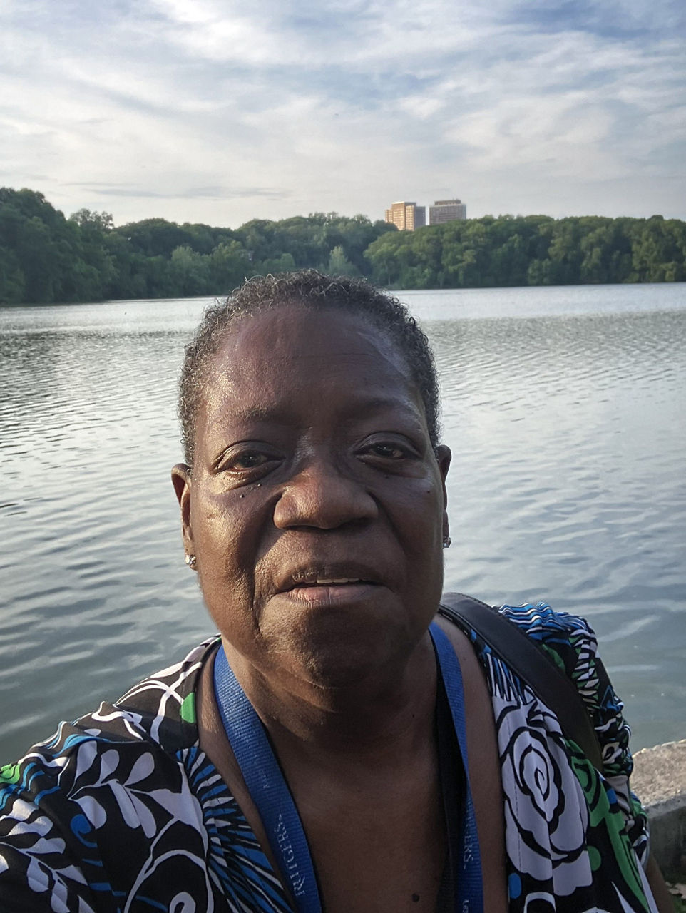 A selfie of Yolanda taken by a lake in a park with trees and a building in the background.