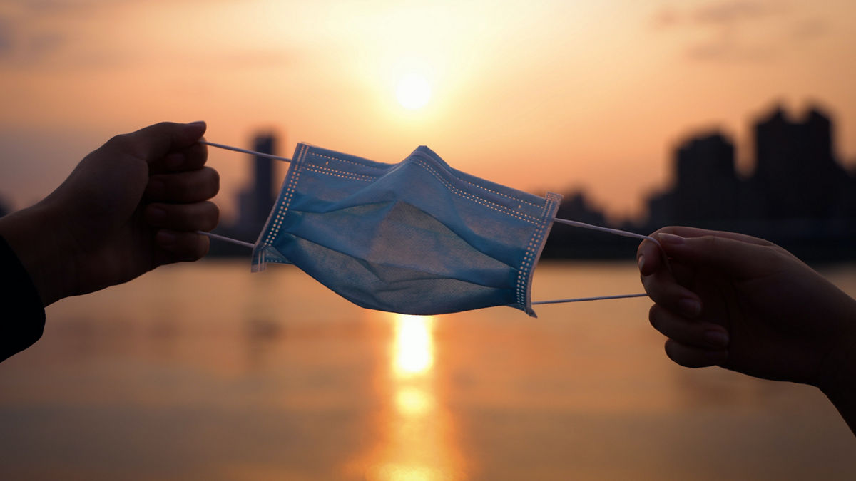 Two hands holding opposite ends of a mask in front of a sunset.