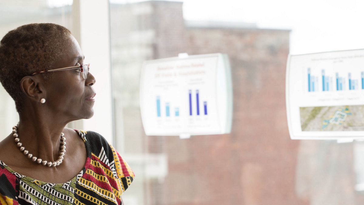 A woman refers to bar charts taped to a window while facilitating a meeting. 