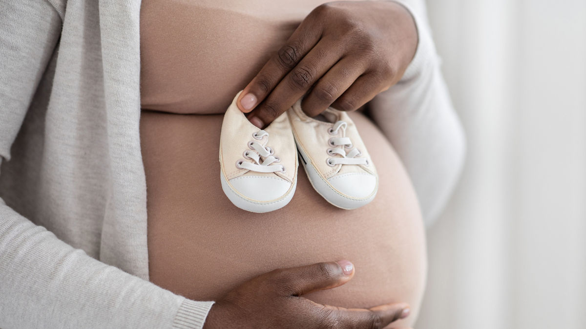 Desired Pregnancy. Black Pregnant Woman Embracing Belly And Holding Small Baby Shoes Near Her Tummy, Expectant Lady Enjoying Maternity Time, Cropped Image With Selective Focus, Closeup Shot