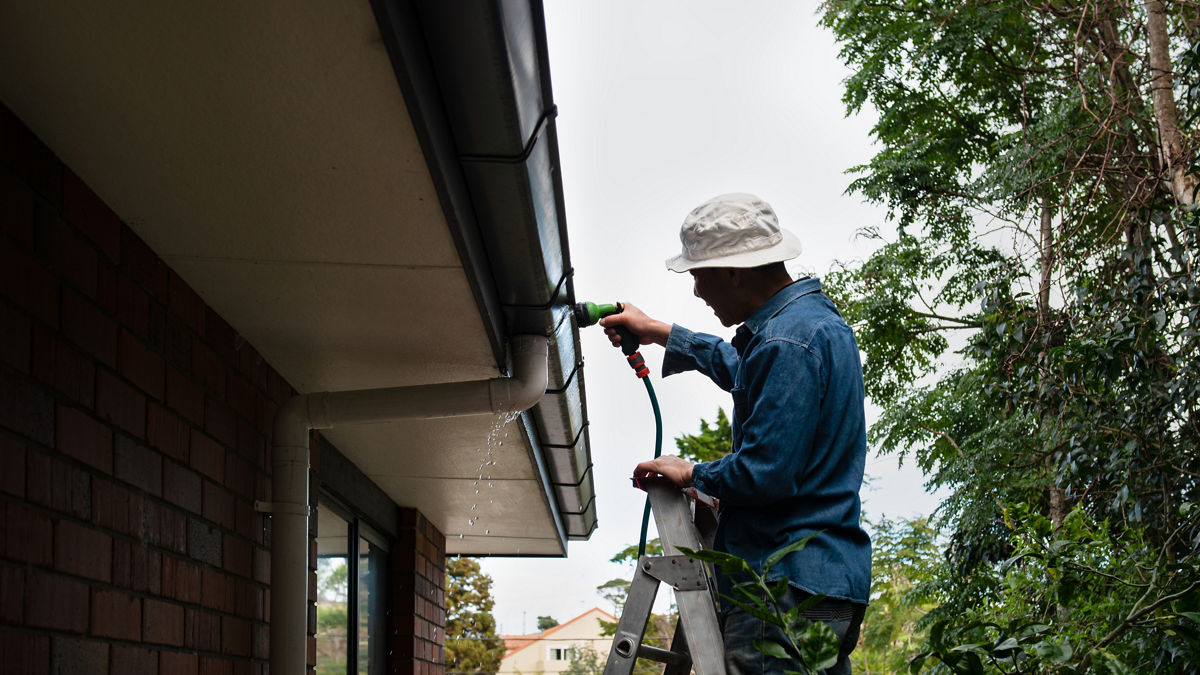 A man in a white bucket hat rinsing gutters with a garden hose while standing atop a ladder.