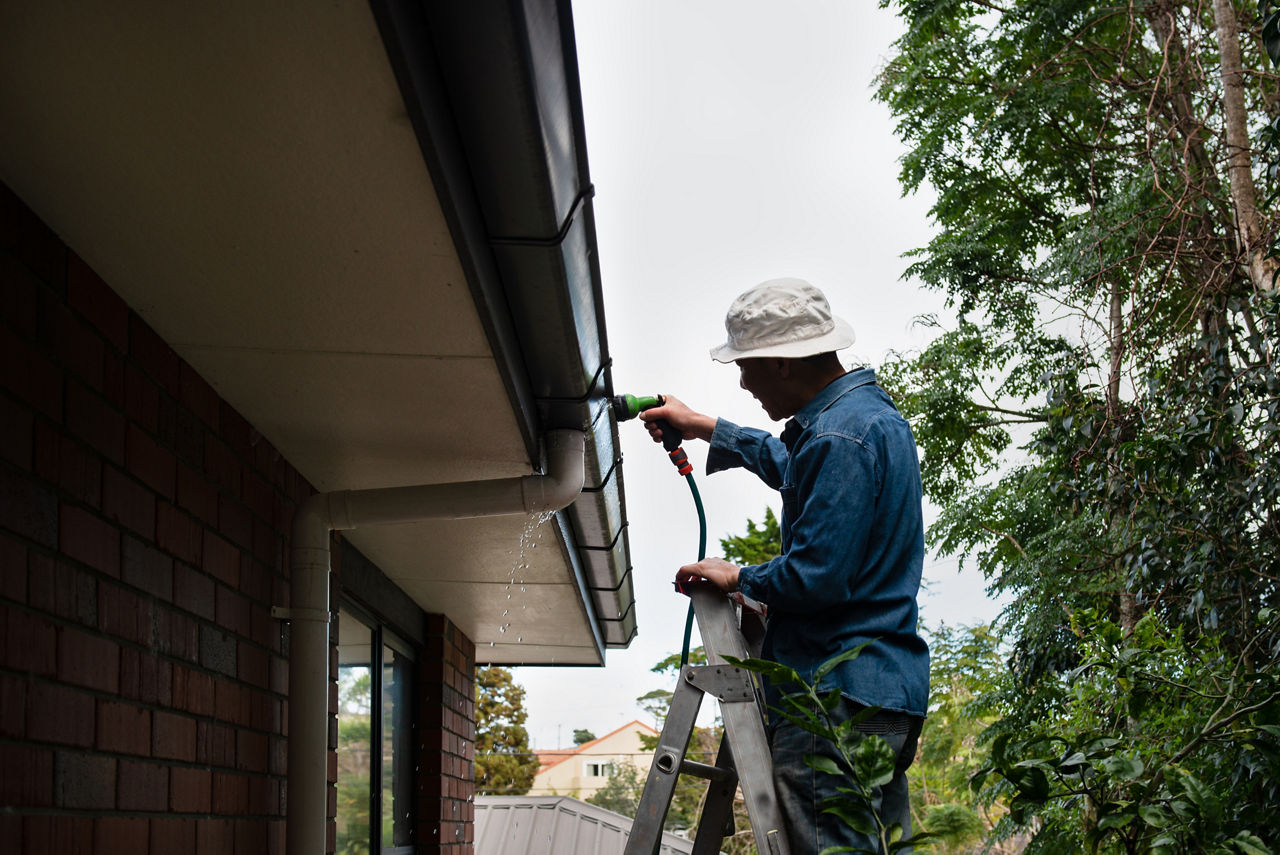 A man in a white bucket hat rinsing gutters with a garden hose while standing atop a ladder.