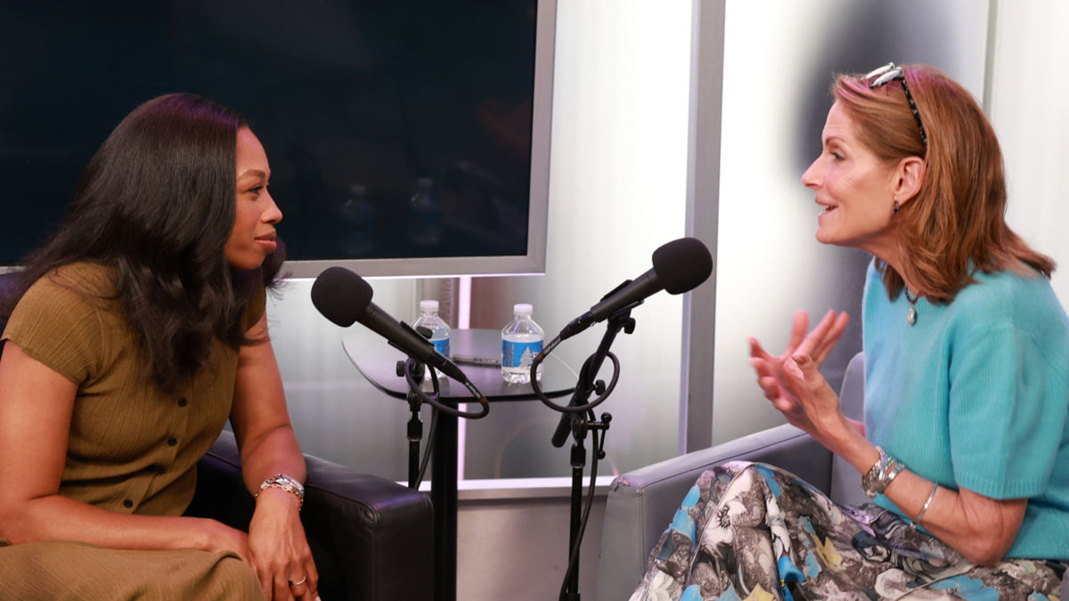 Two people seated across from each other in a recording studio, speaking into microphones during a conversation.