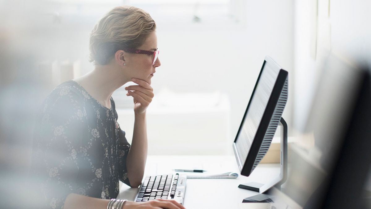 One white businesswoman working in office at desktop computer, focused, eyeglasses, chin on hand, sitting.