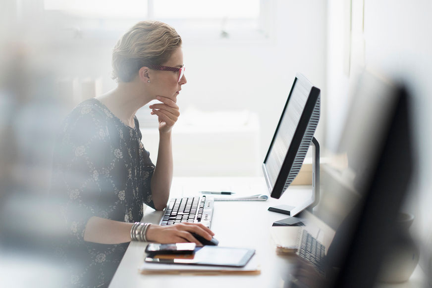 One white businesswoman working in office at desktop computer, focused, eyeglasses, chin on hand, sitting.