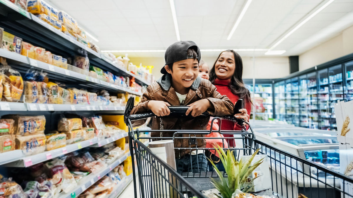 A young boy having fun on a shopping cart while out buying groceries with his mother.