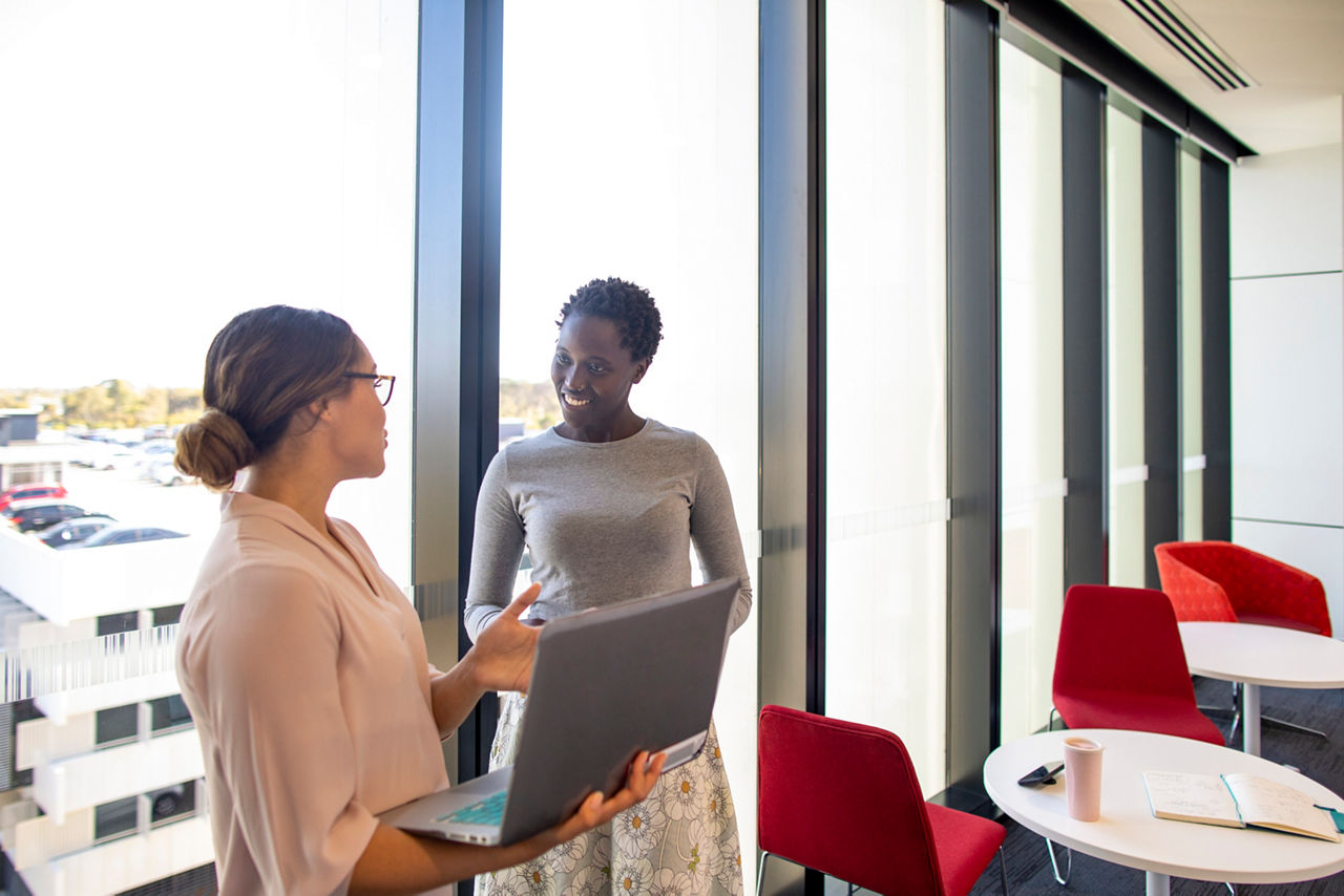 Two women conversing in front a large office window.