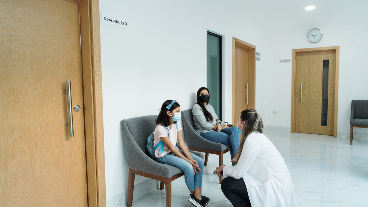 A doctor with a face mask talking to a girl patient and her mother at a clinic.